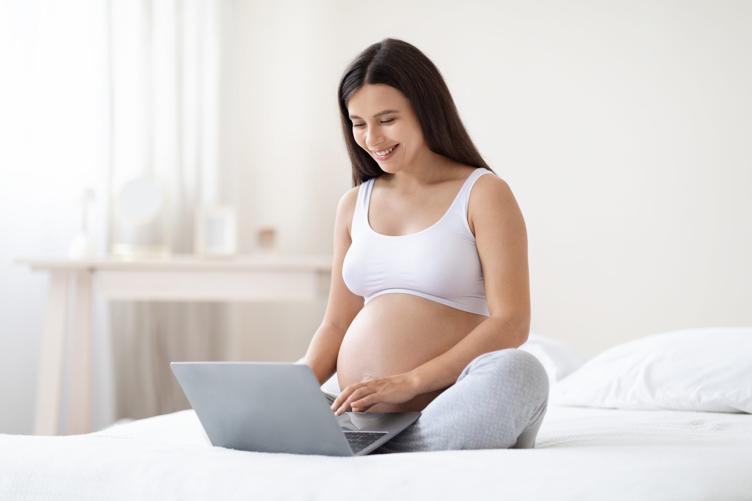 Happy attractive young pregnant woman using computer at home, cheerful expecting lady in homewear sitting on bed, websurfing, shopping on Internet, choosing baby clothes, copy space